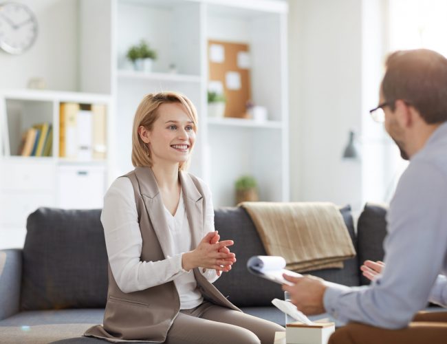 Young blonde smiling woman sitting on couch and looking at counselor during discussion of ways of solving problem